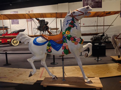 A beautiful hand-carved, painted antique carousel horse awaits installation, near a historic plane and automobile.